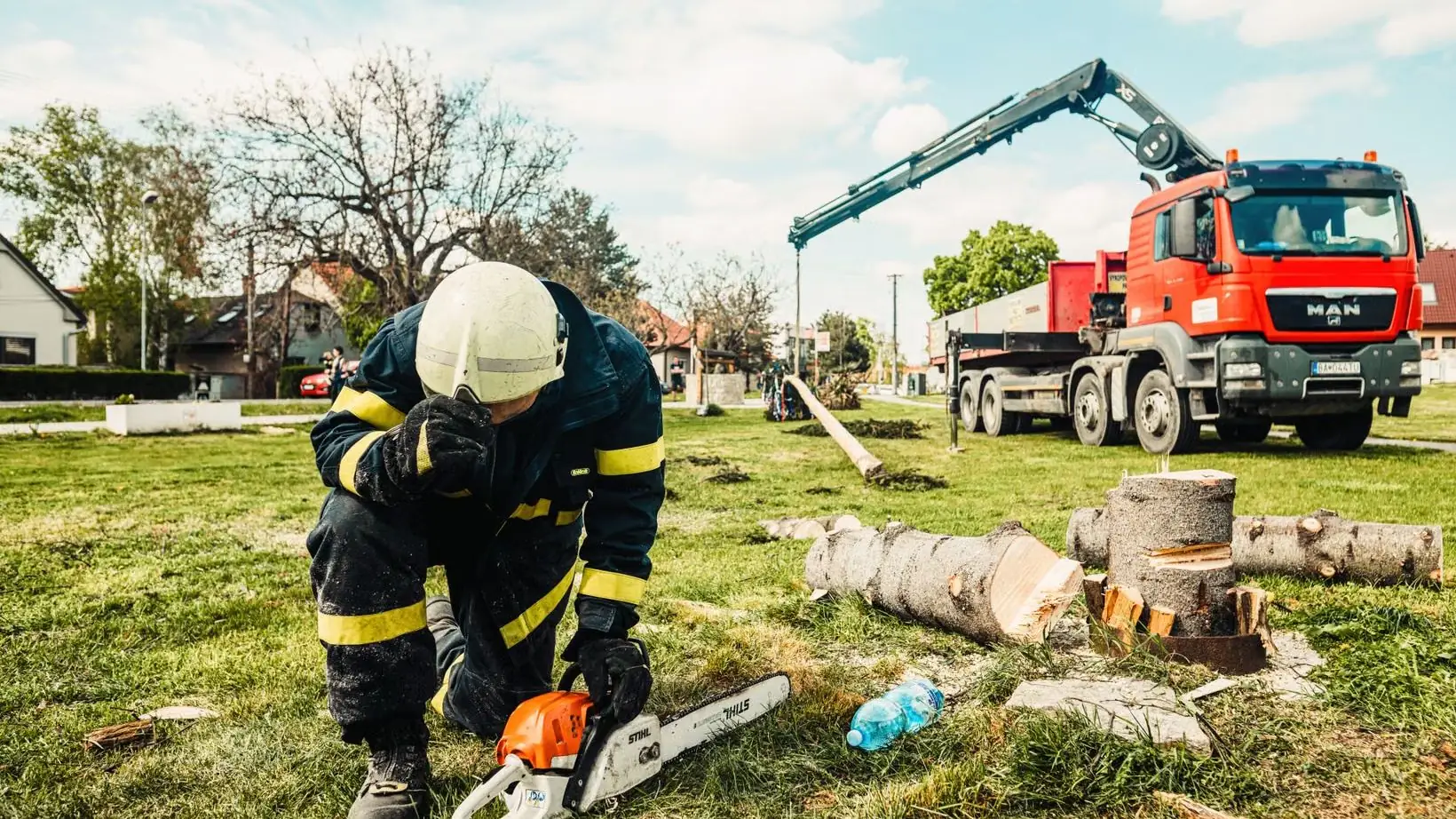 A worker on the ground with a chainsaw beside sectioned trunk logs and a crane truck
